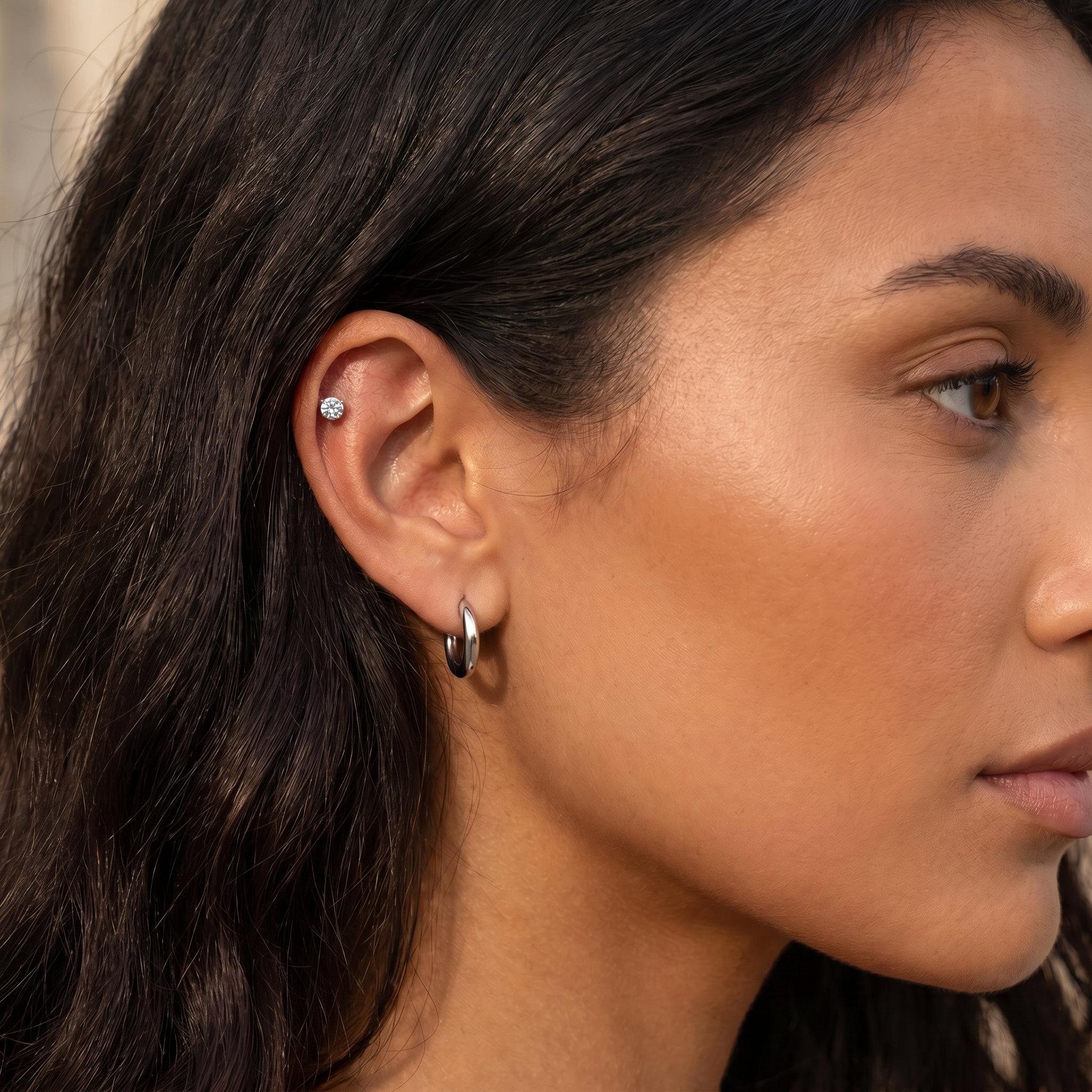 Close-up of a woman's ear showcasing two silver hoop earrings, with one small silver stud earring on the upper ear cartilage.