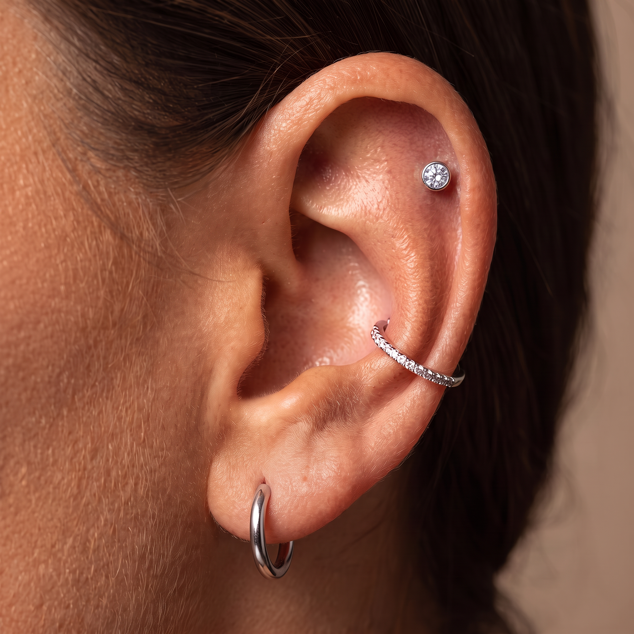 Close-up of a person's ear showcasing multiple silver piercings: a hoop earring in the lobe, a curved cartilage hoop with small clear stones, and a round stud in the lobe.