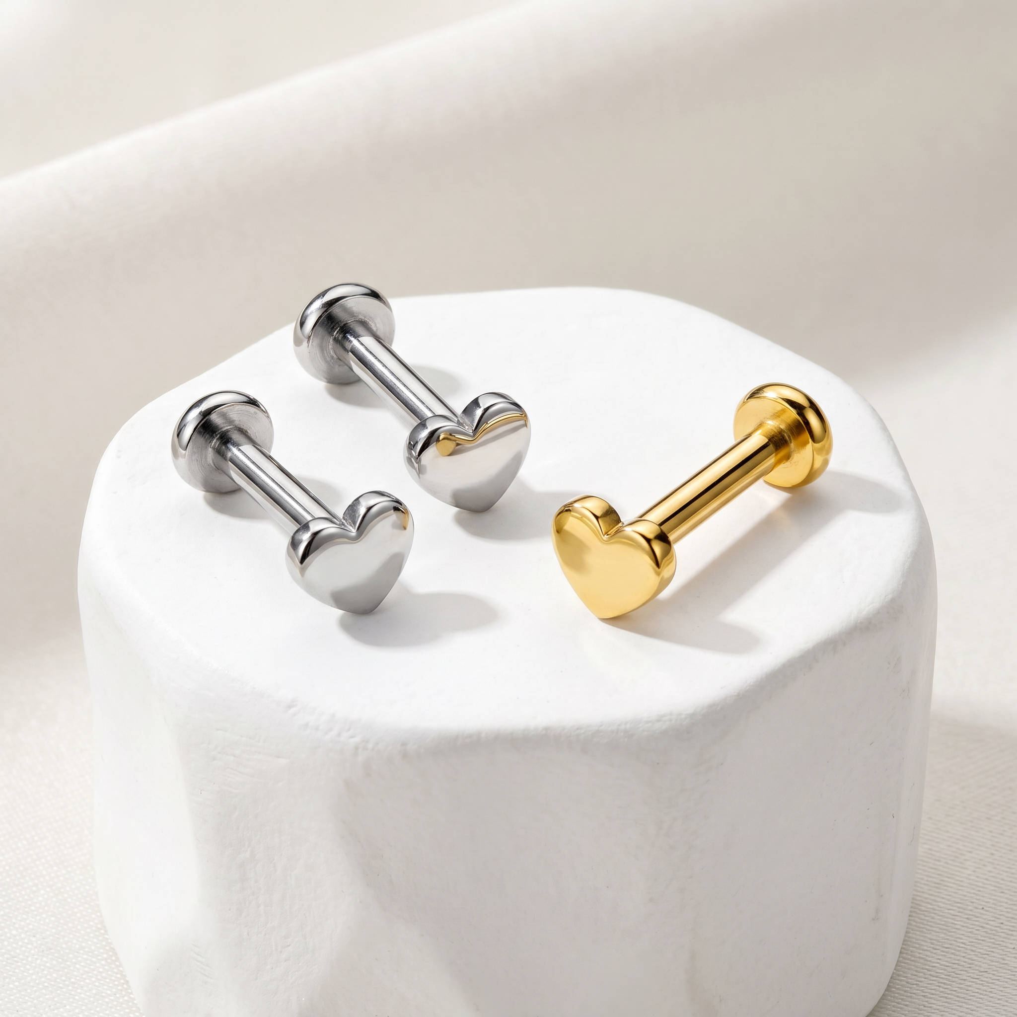 Three heart-shaped barbell stud earrings displayed on a white cylindrical pedestal. Two are silver-toned and one is gold-toned, set against a soft beige fabric background.