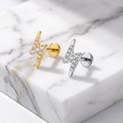 Two lightning bolt stud earrings on a white marble slab, one in yellow gold with clear stones and one in silver with clear stones.