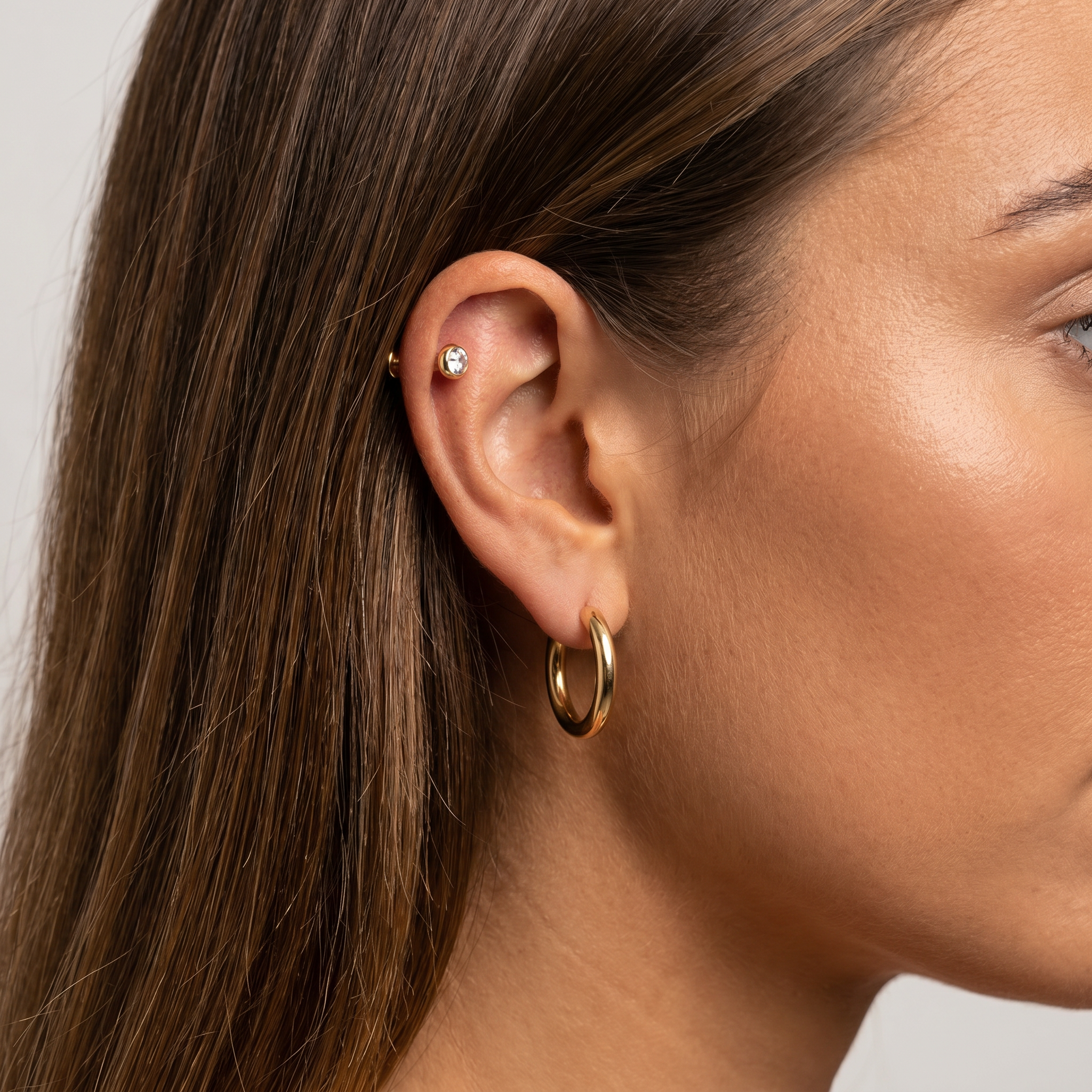 A close-up of a woman's ear wearing a polished gold hoop earring and a small gold stud with a clear gemstone, set against a neutral light gray background.