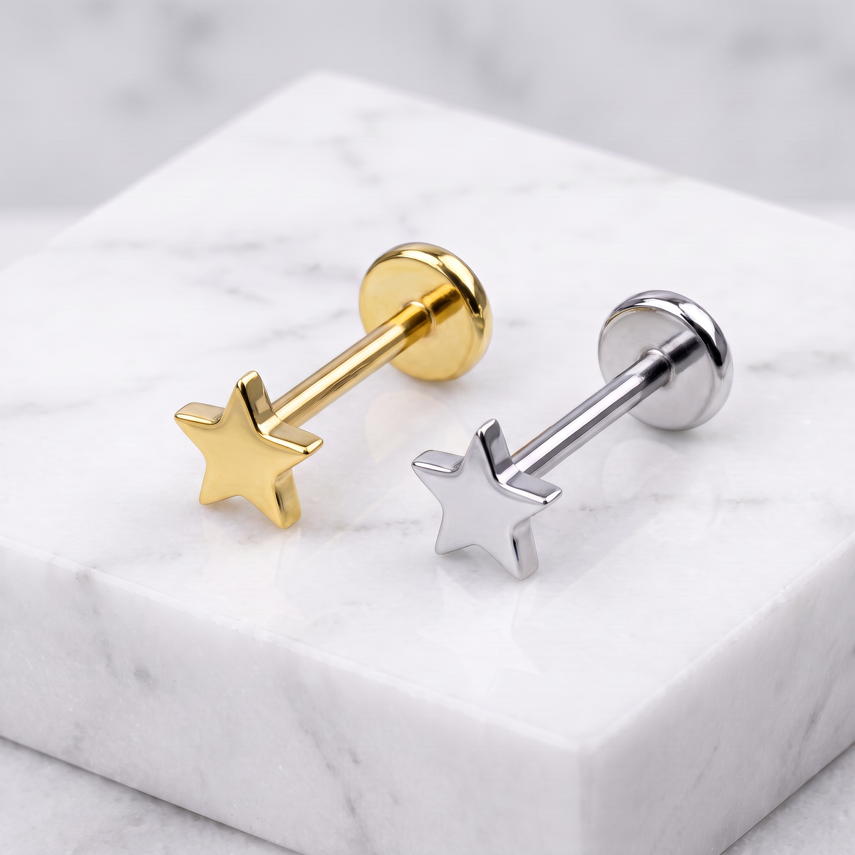 Two star-shaped stud earrings, one in polished gold and one in polished silver, displayed on a white marble block.
