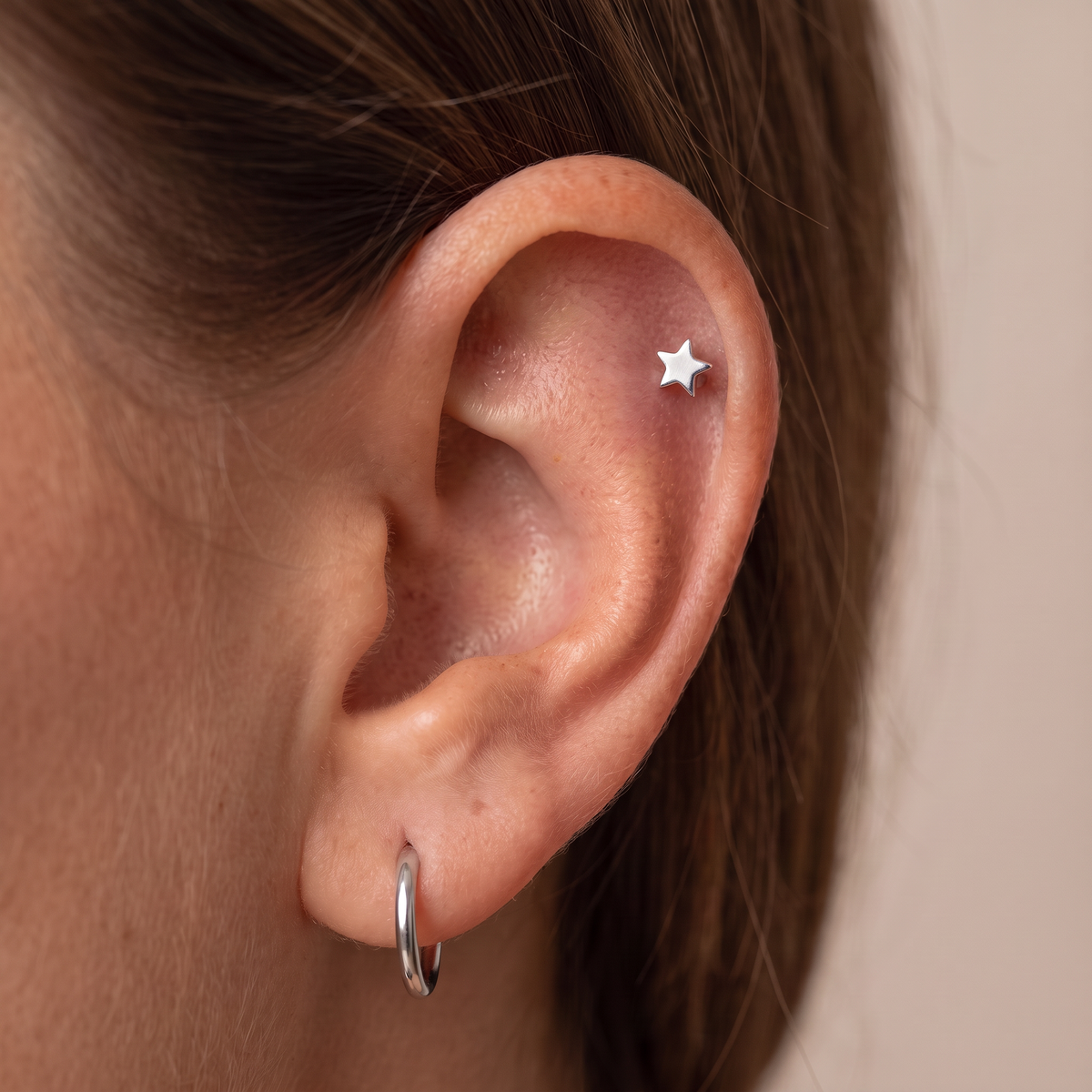 A close-up of a person's ear showing a small silver star stud earring in the cartilage and a silver hoop earring in the lobe, against a soft beige background.