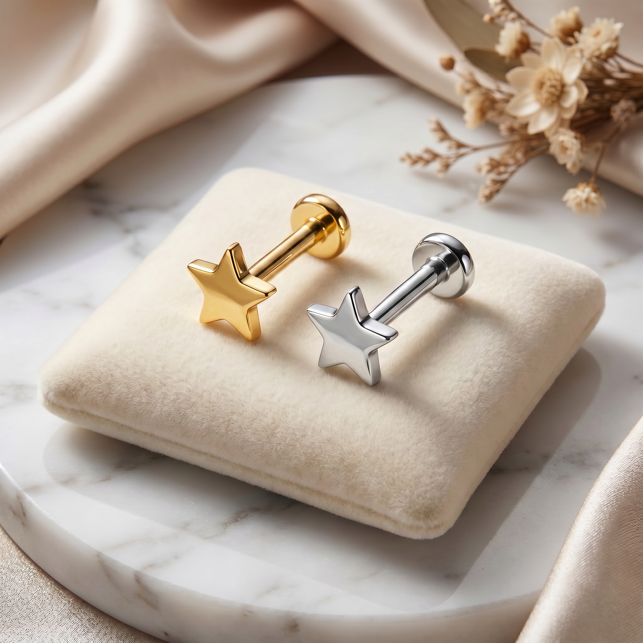 Two star-shaped stud earrings, one gold-toned and one silver-toned, displayed on a cream velvet jewelry cushion over a white marble surface with draped satin fabric and dried flowers in the background.
