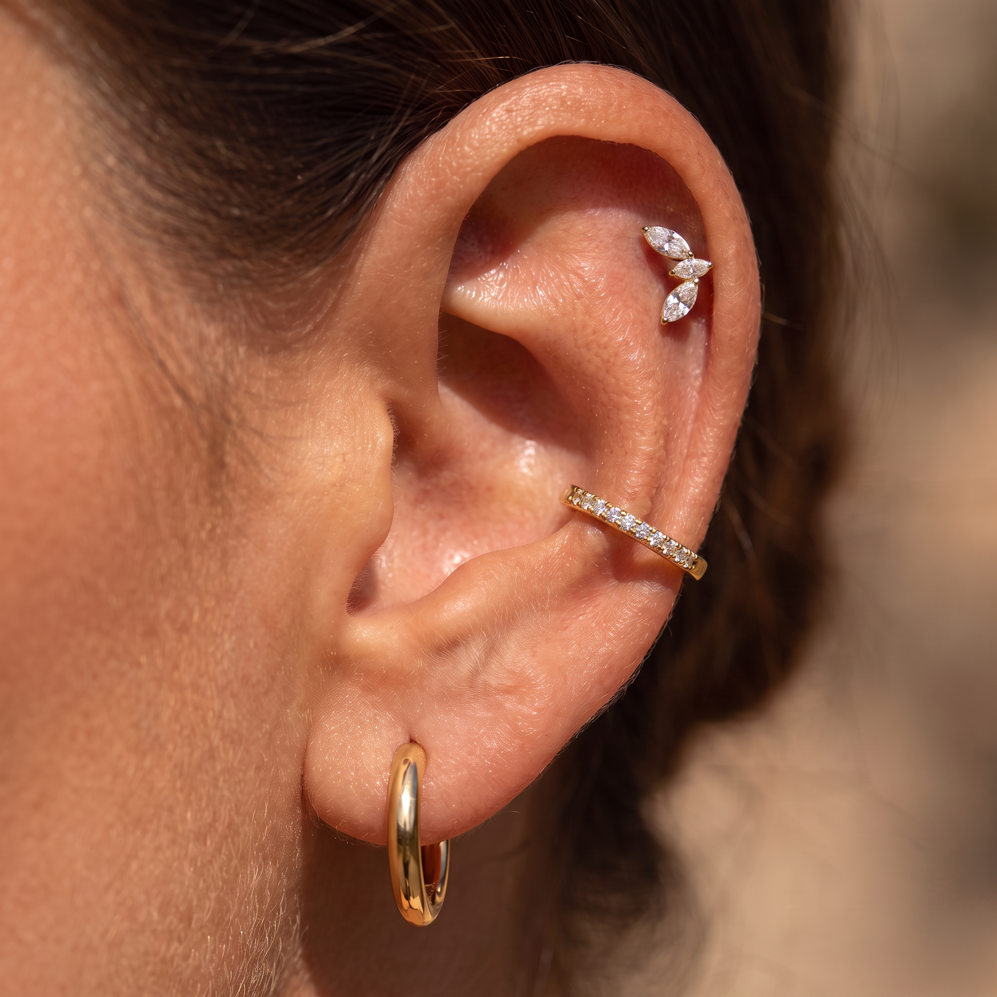 Close-up of a person's ear showcasing three gold ear piercings: a small hoop earring in the cartilage, a curved pavé band in the helix, and a butterfly-shaped stud in the lobe.