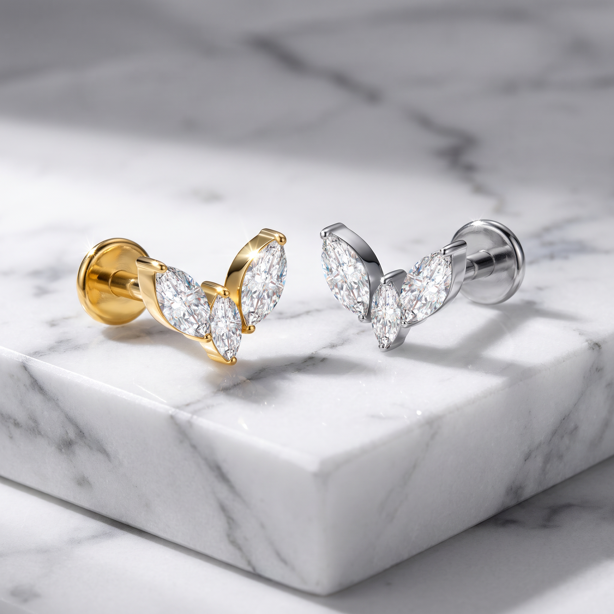 Two butterfly-shaped stud earrings with marquise-cut clear stones, one in yellow gold and one in silver, displayed on a white marble block.