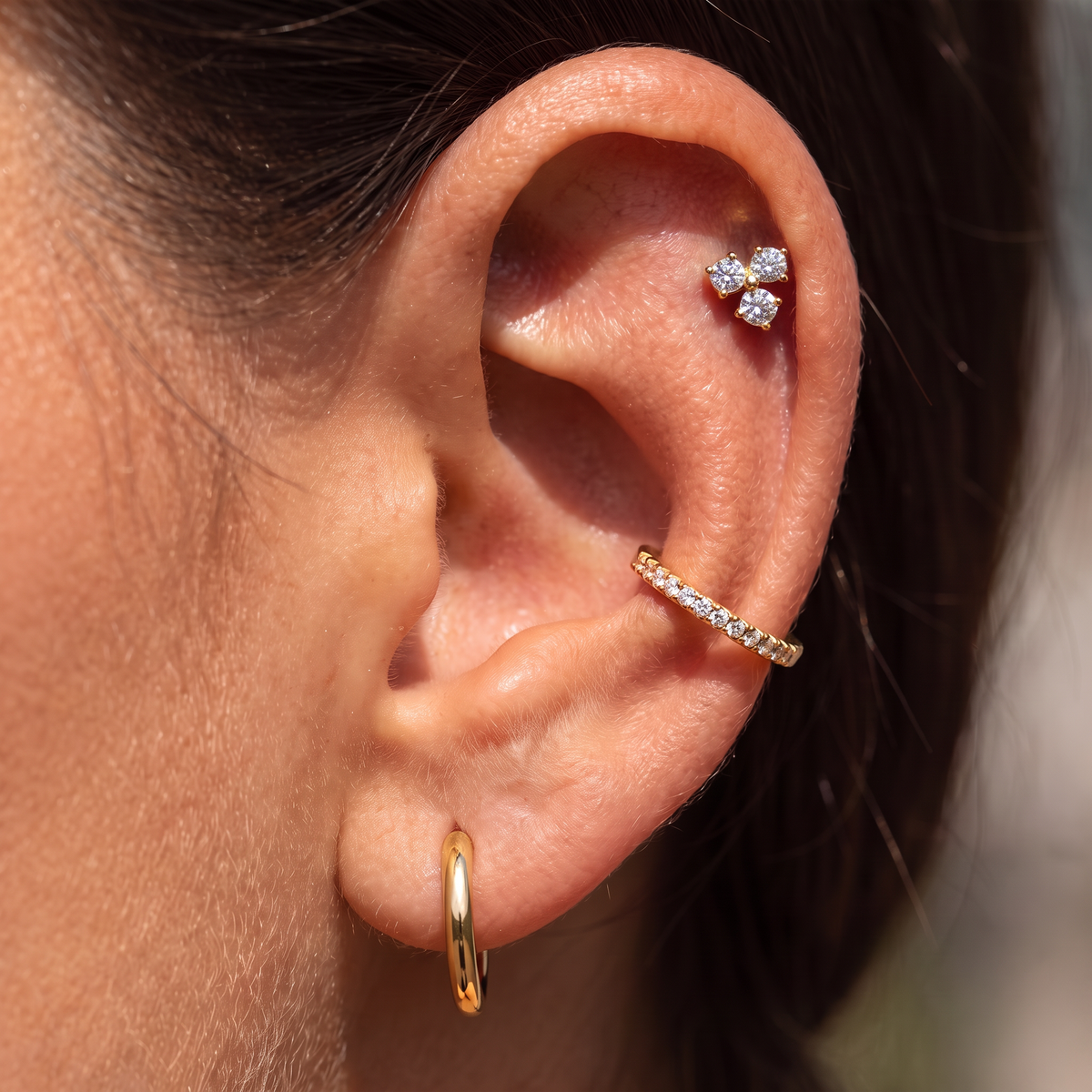 Close-up of a person's ear showcasing three gold and silver stud earrings, including a small bow-shaped silver earring, a silver pavé cartilage hoop, and a gold huggie hoop.