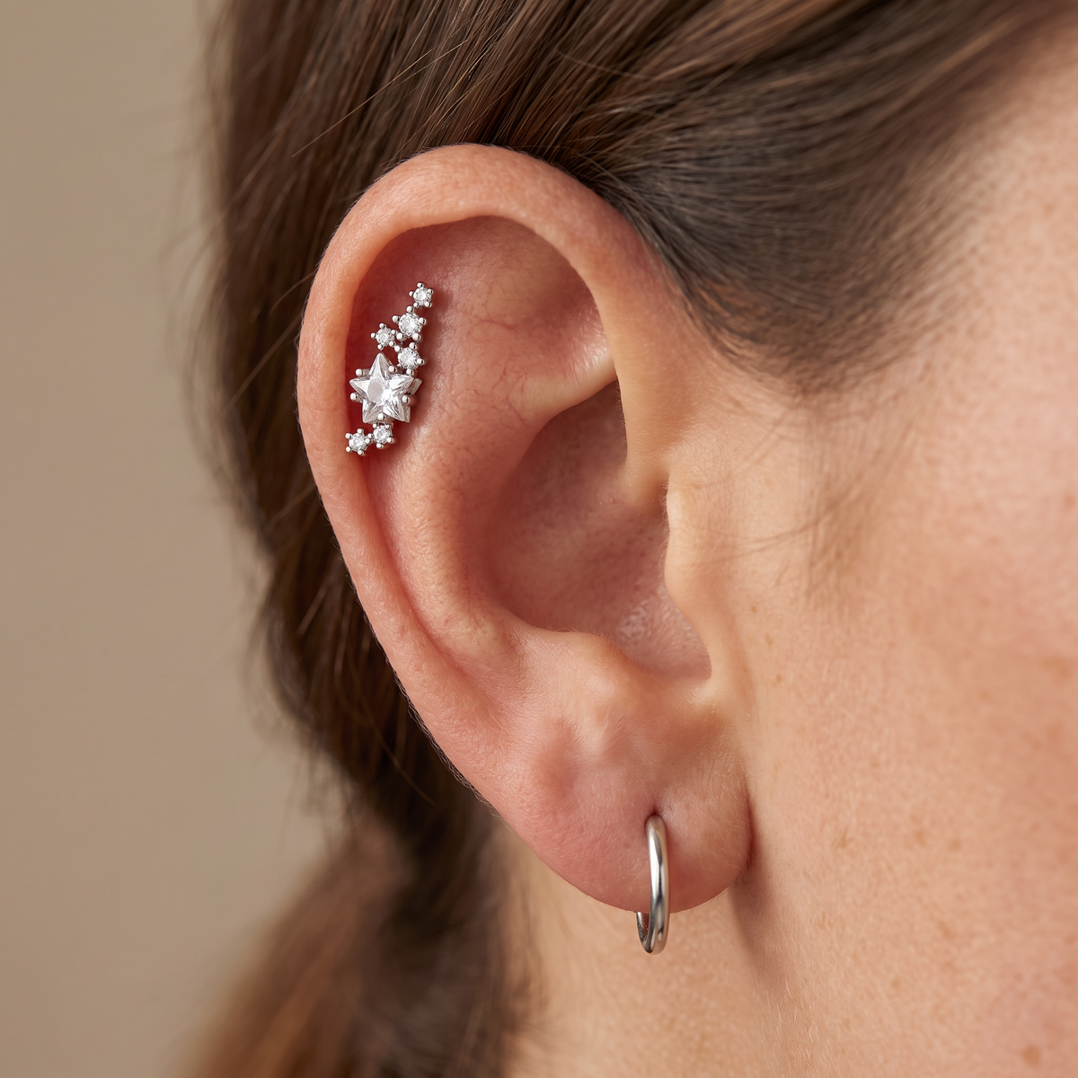 A close-up of a person's ear showcasing a silver-toned cluster ear cuff with clear stones, paired with a simple silver hoop earring.