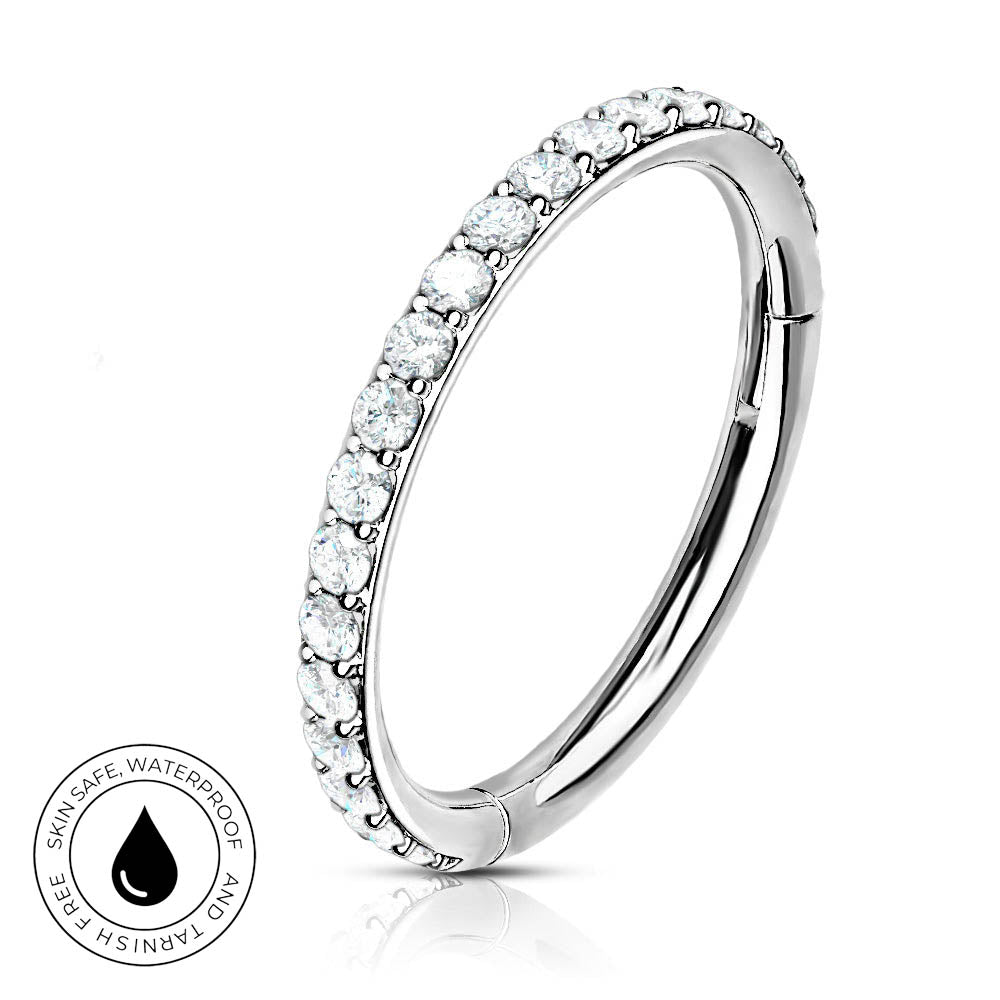 A silver-toned curved ring band with a row of clear round gemstones along the front, shown on a white background with a small circular 'skin-safe waterproof' badge in the lower left.
