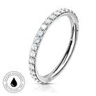 A silver-toned curved ring band with a row of clear round gemstones along the front, shown on a white background with a small circular 'skin-safe waterproof' badge in the lower left.