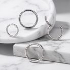 Four silver-toned hoop and C-shaped body jewelry pieces arranged on white marble blocks and a marble tray.