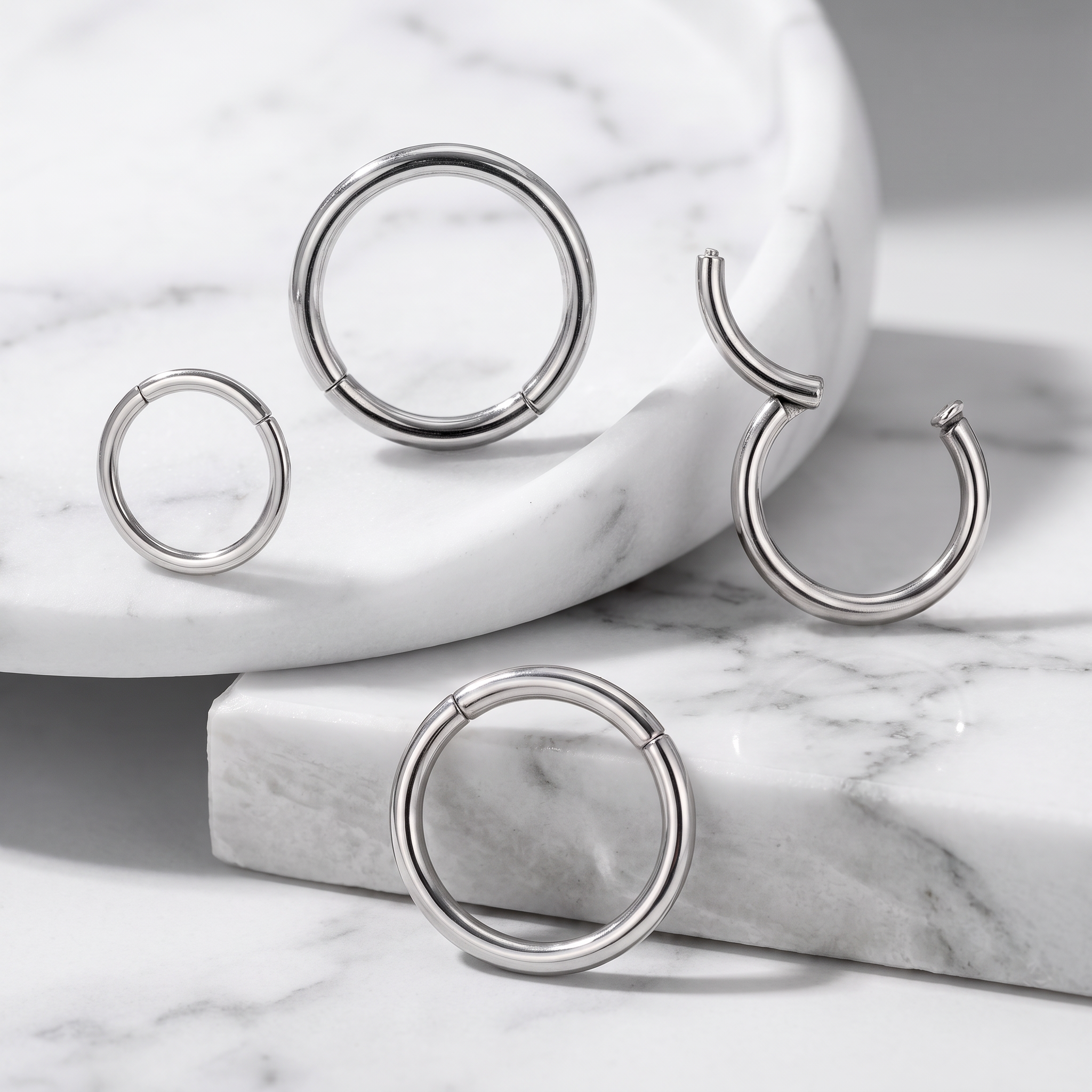 Four silver-toned hoop and C-shaped body jewelry pieces arranged on white marble blocks and a marble tray.