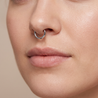 A close-up of a person's face wearing a thin silver metal nose hoop ring on the left nostril, set against a soft beige background.