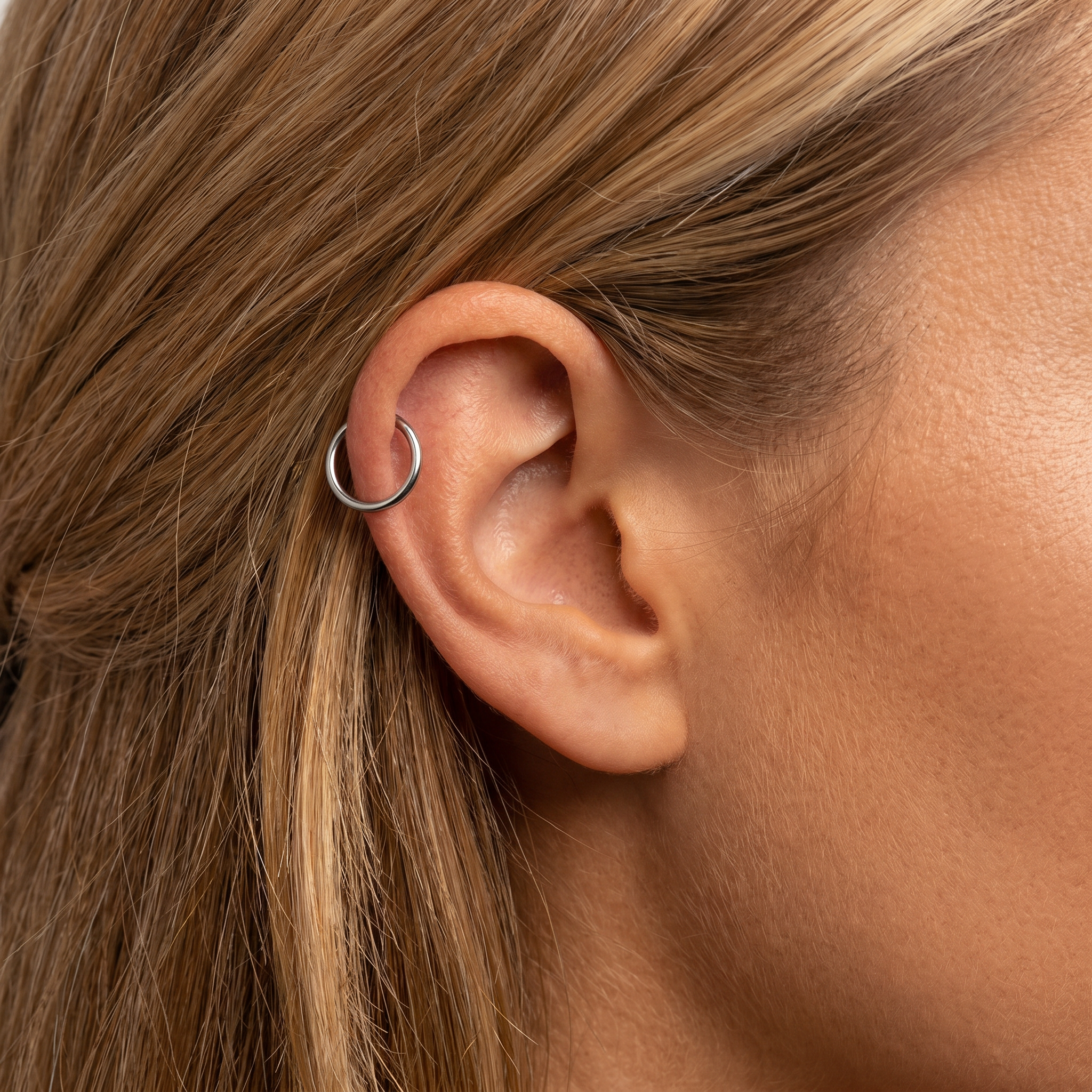 A close-up of a person's ear wearing a small silver hoop earring in the helix position, with light brown hair and skin visible.