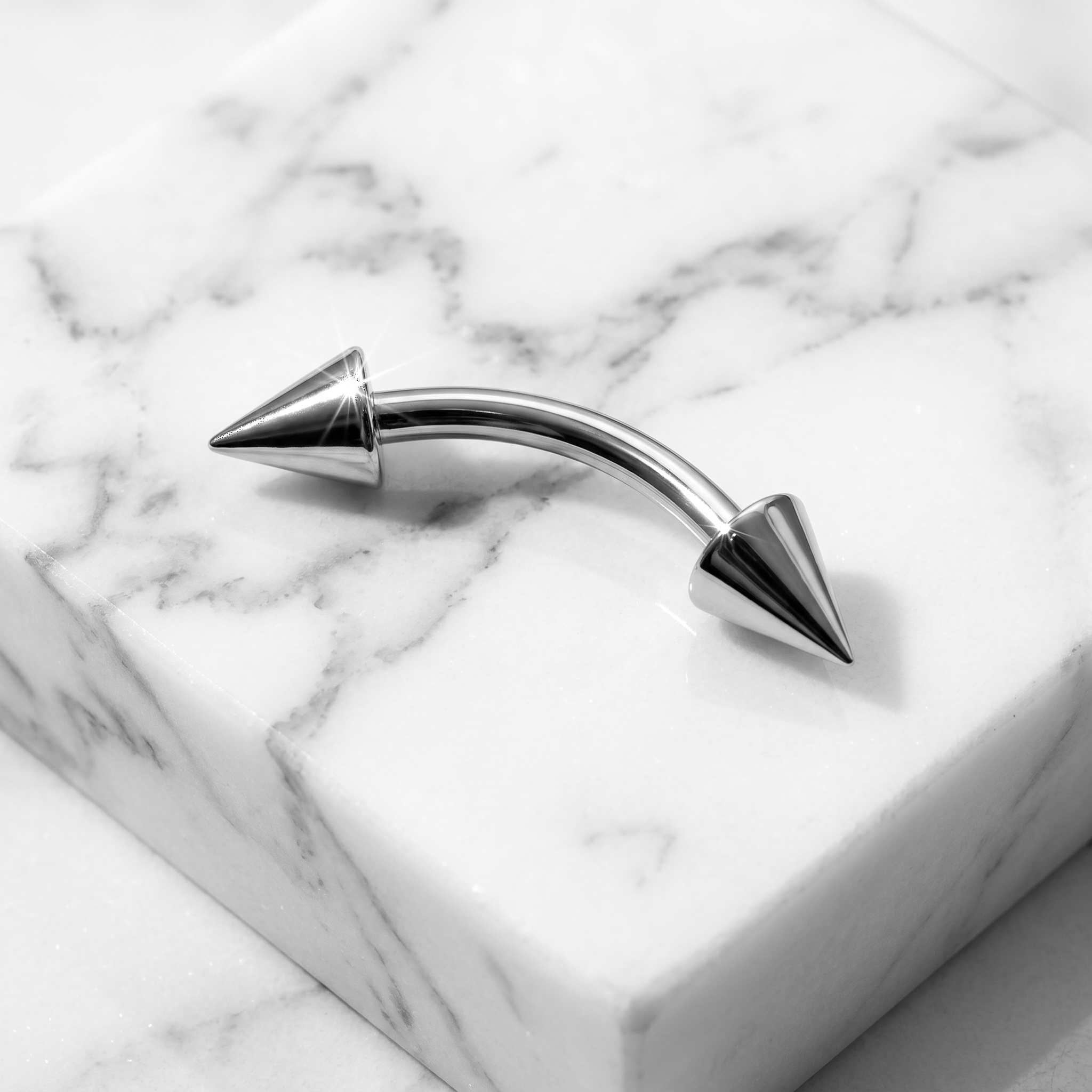 A curved barbell piercing with metallic silver conical ends, resting on a white and gray marble block.