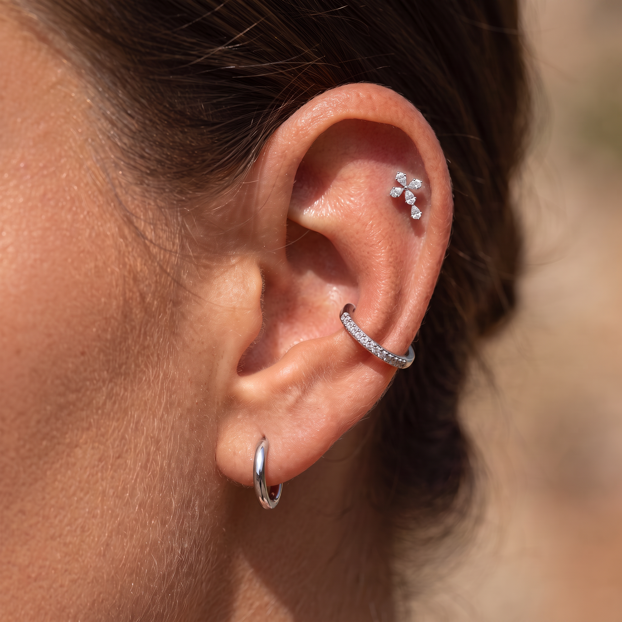 Close-up of a person's ear showcasing three silver ear piercings: a small hoop earring, a curved cartilage hoop adorned with clear crystals, and a small floral cluster stud on the upper ear cartilage.