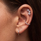 Close-up of a person's ear showcasing silver ear piercings: a small hoop in the lobe, a curved ear cuff with clear stones, and a cluster of three flower-shaped ear studs.