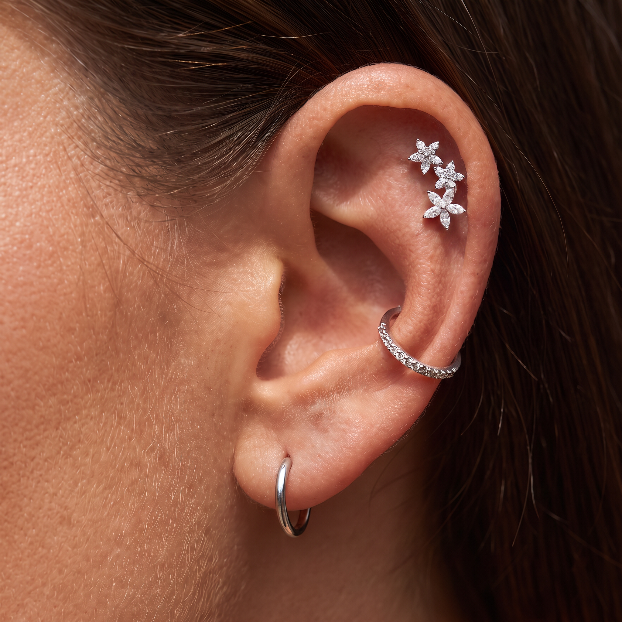 Close-up of a person's ear showcasing silver ear piercings: a small hoop in the lobe, a curved ear cuff with clear stones, and a cluster of three flower-shaped ear studs.
