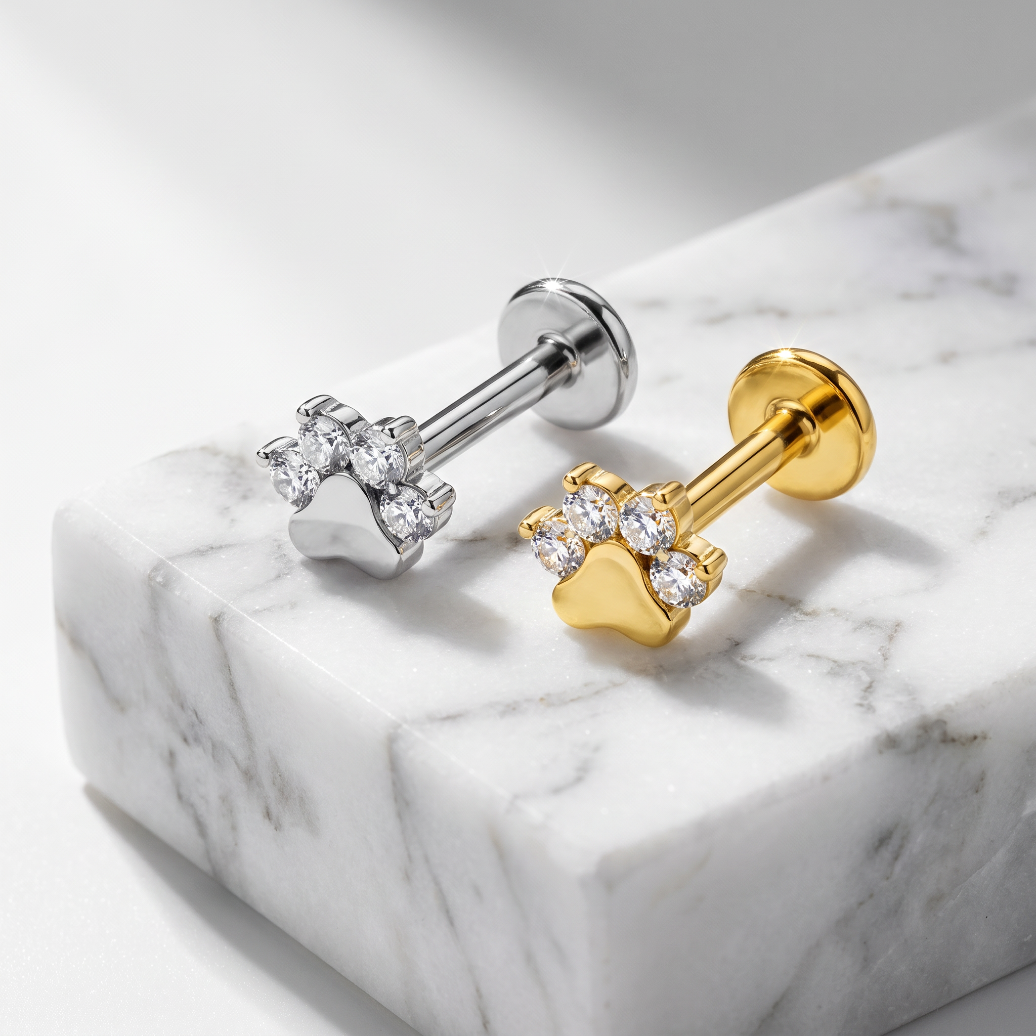 Two stud earrings displayed on a white marble block. One earring is silver-toned with a paw-shaped setting set with clear stones, and the other is gold-toned with a matching paw-shaped setting and clear stones.