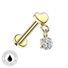 Gold barbell lobe piercing stud with a heart-shaped top and a dangling round clear gemstone charm, shown on a white background with a small skin-safe waterproof badge in the lower-left corner.
