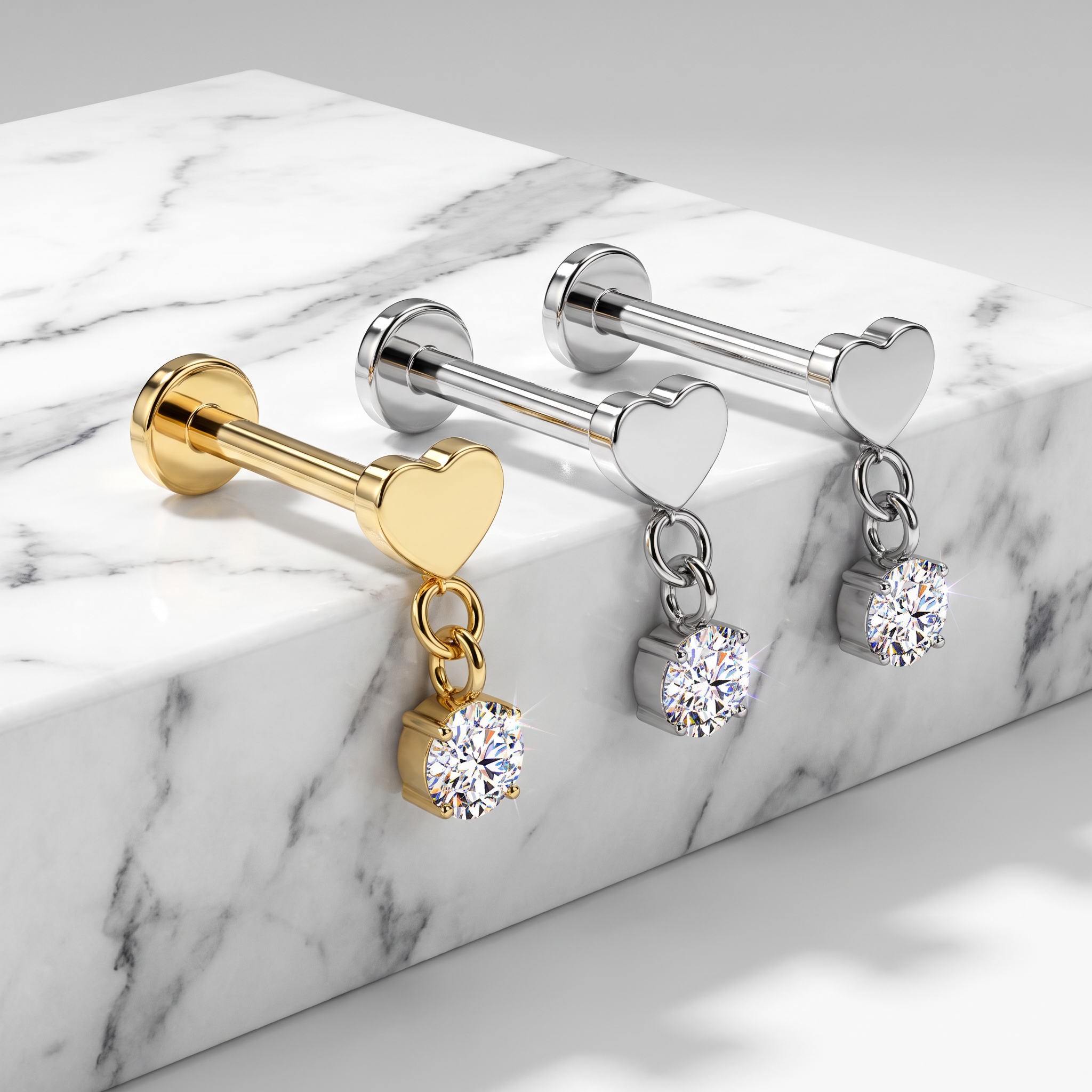 Three heart-shaped barbell stud earrings with dangling round-cut clear gemstones are arranged on a white marble block. One earring is gold-toned, while the other two are silver-toned.