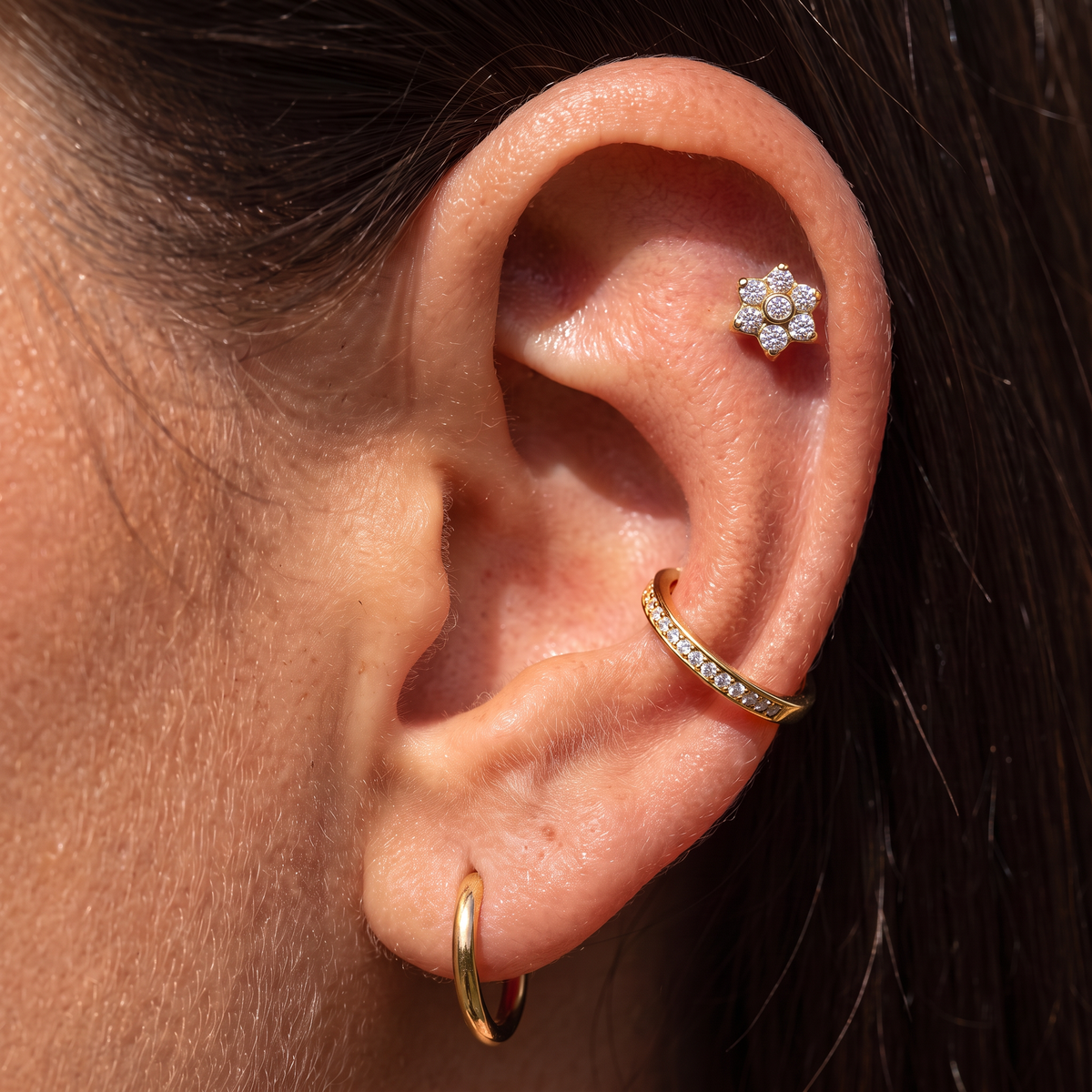 Close-up of a person's ear showcasing three gold ear piercings: a small flower-shaped stud with clear stones on the helix, a curved cartilage hoop with clear stones, and a simple gold hoop in the lobe. Dark hair frames the ear.