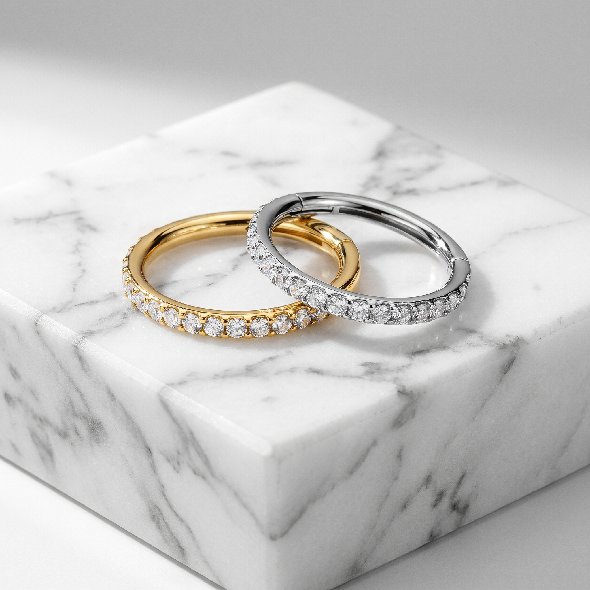 Two slim hoop rings displayed on a white marble block. One ring is yellow gold with a row of clear stones, and the other is silver-toned with a row of clear stones.