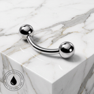 A curved barbell piercing jewelry piece with two polished silver ball ends, displayed on a white marble block. A circular 'SkinCafe Waterproof' logo is visible in the bottom-left corner.
