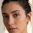 Close-up of a woman's face showing three small silver geometric eyebrow piercings on her brow, with her hair pulled back and a soft neutral background.