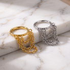 Two statement rings with pyramid-accented bands and draped chain details, shown in gold and silver finishes on a white marble surface.