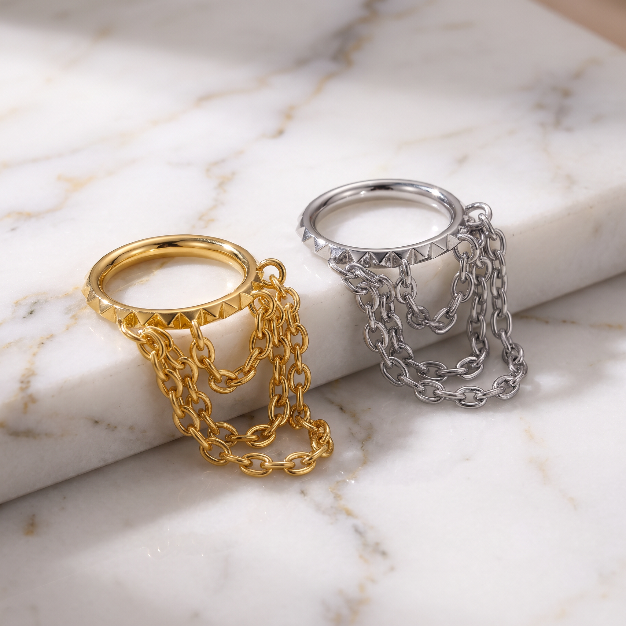Two statement rings with pyramid-accented bands and draped chain details, shown in gold and silver finishes on a white marble surface.