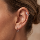 Close-up of a person's ear wearing a silver hoop earring in the lobe and a silver curved ear cuff set with small clear stones, against a soft beige background.