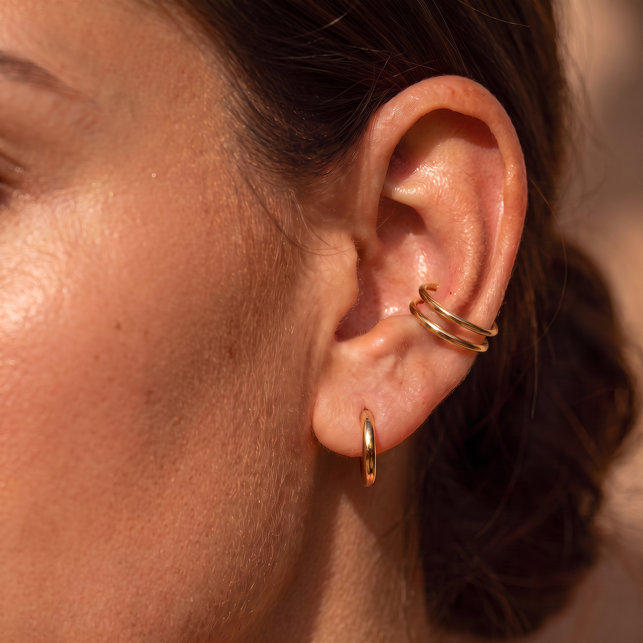 A close-up of a person's ear showcasing gold body jewelry, including a small hoop earring, a double-hoop cartilage piercing, and a curved helix piercing.