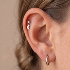 Close-up of a person's ear showcasing a silver crescent moon and diamond stud earring in the cartilage, with a silver hoop earring in the lobe, against a neutral beige background.