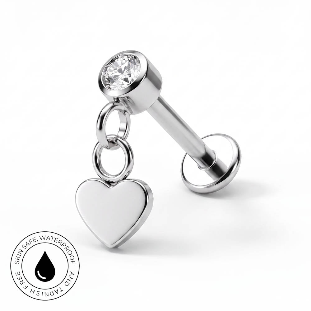 A silver-toned labret piercing stud with a clear round gemstone and a dangling heart charm, shown on a white background with a small circular skin-safe waterproof badge in the lower left corner.