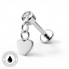 A silver-toned labret piercing stud with a clear round gemstone and a dangling heart charm, shown on a white background with a small circular skin-safe waterproof badge in the lower left corner.
