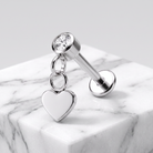 A silver-toned labret piercing stud with a clear round gemstone and a dangling polished heart charm, displayed on a white marble block against a light gray background.
