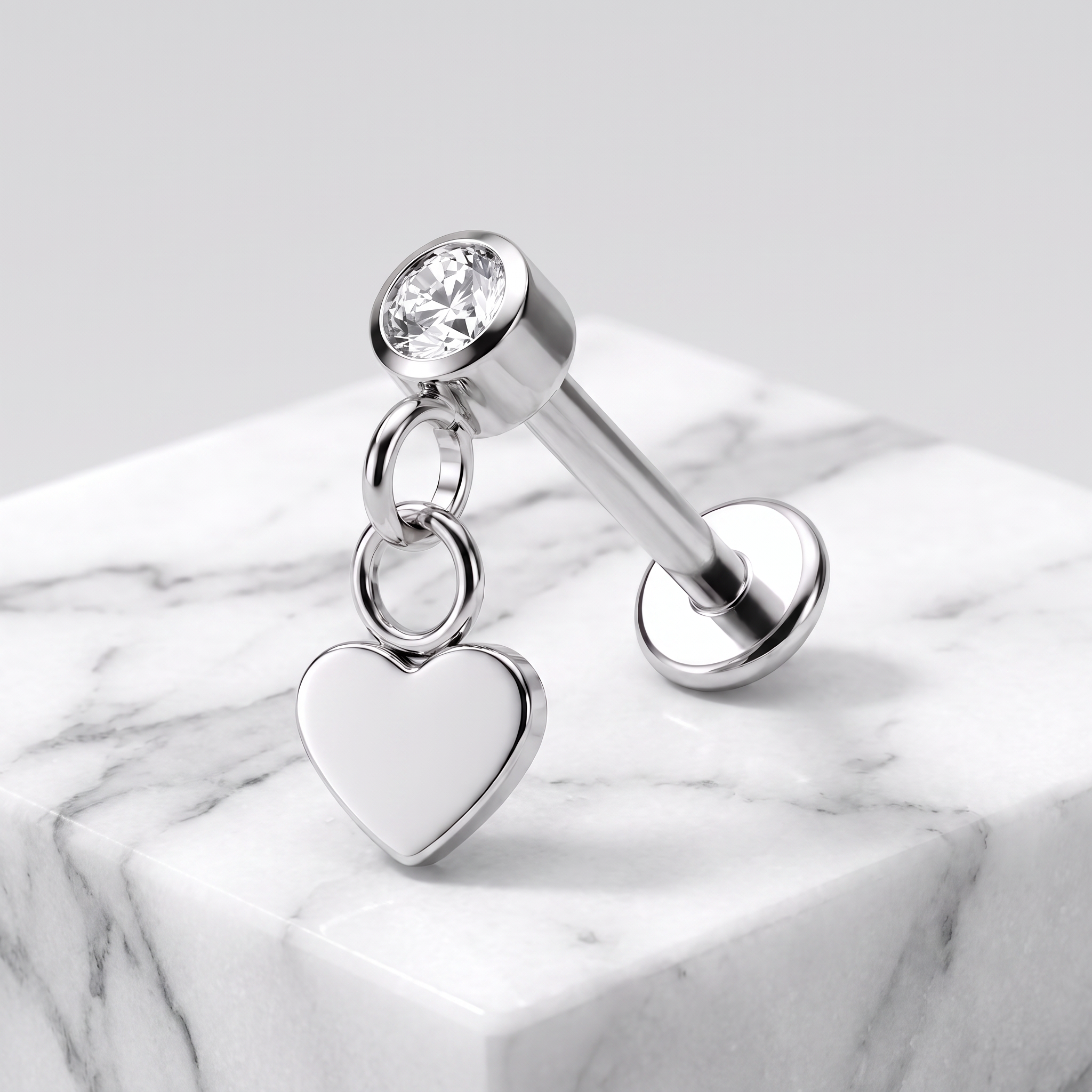 A silver-toned labret piercing stud with a clear round gemstone and a dangling polished heart charm, displayed on a white marble block against a light gray background.