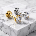Two stud earrings displayed on a white marble block. One earring features a yellow gold post and bezel with a clear round gemstone, while the other features a silver-toned post and bezel with a matching clear gemstone.