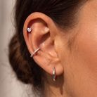 Close-up of a person's ear showcasing multiple silver-toned ear piercings: a small heart-shaped stud in the lobe, a curved ear cuff with small clear stones, and a simple hoop earring in the cartilage.
