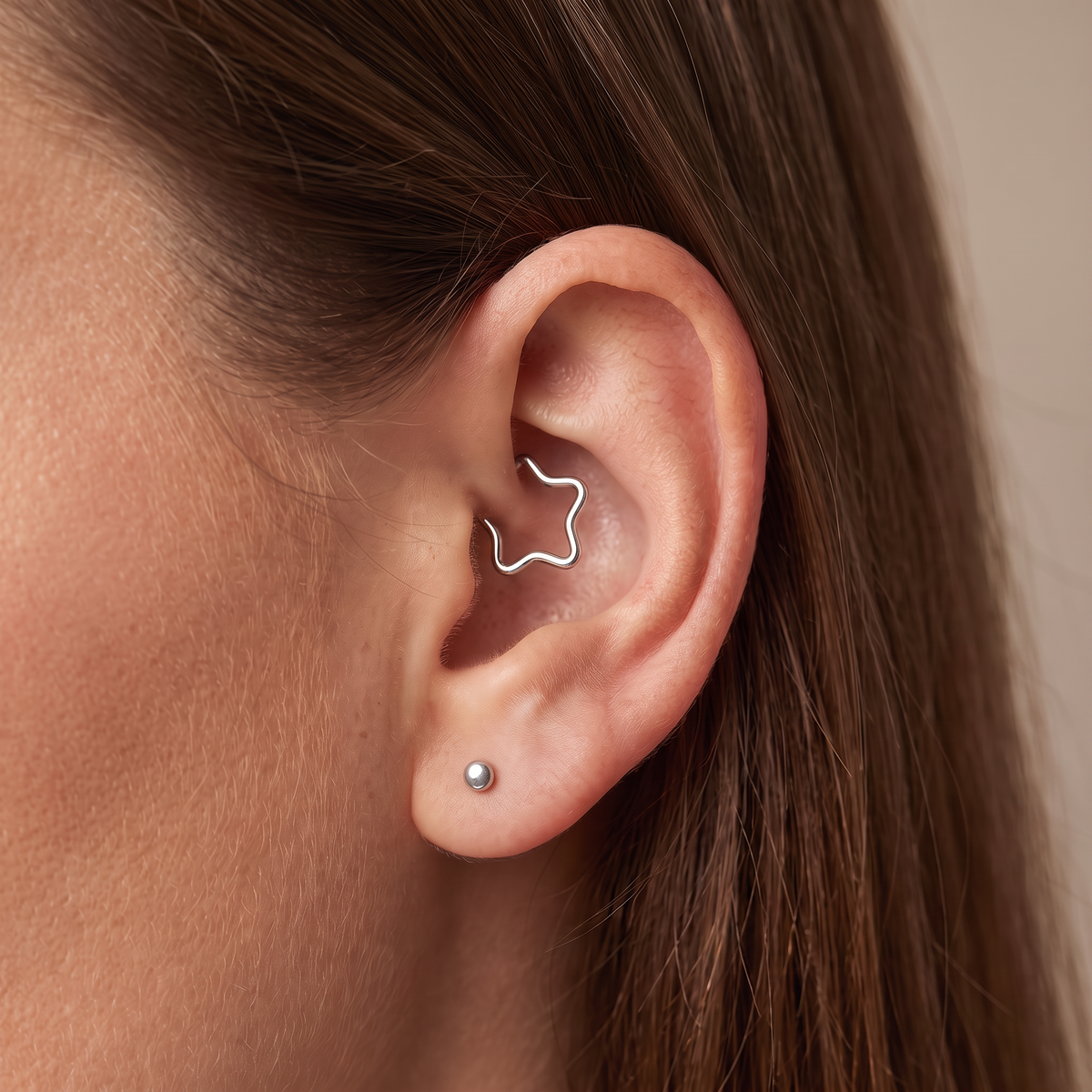 Close-up of a person's ear wearing a delicate silver hoop with a wavy outline and a small silver stud earring, against a soft beige background.