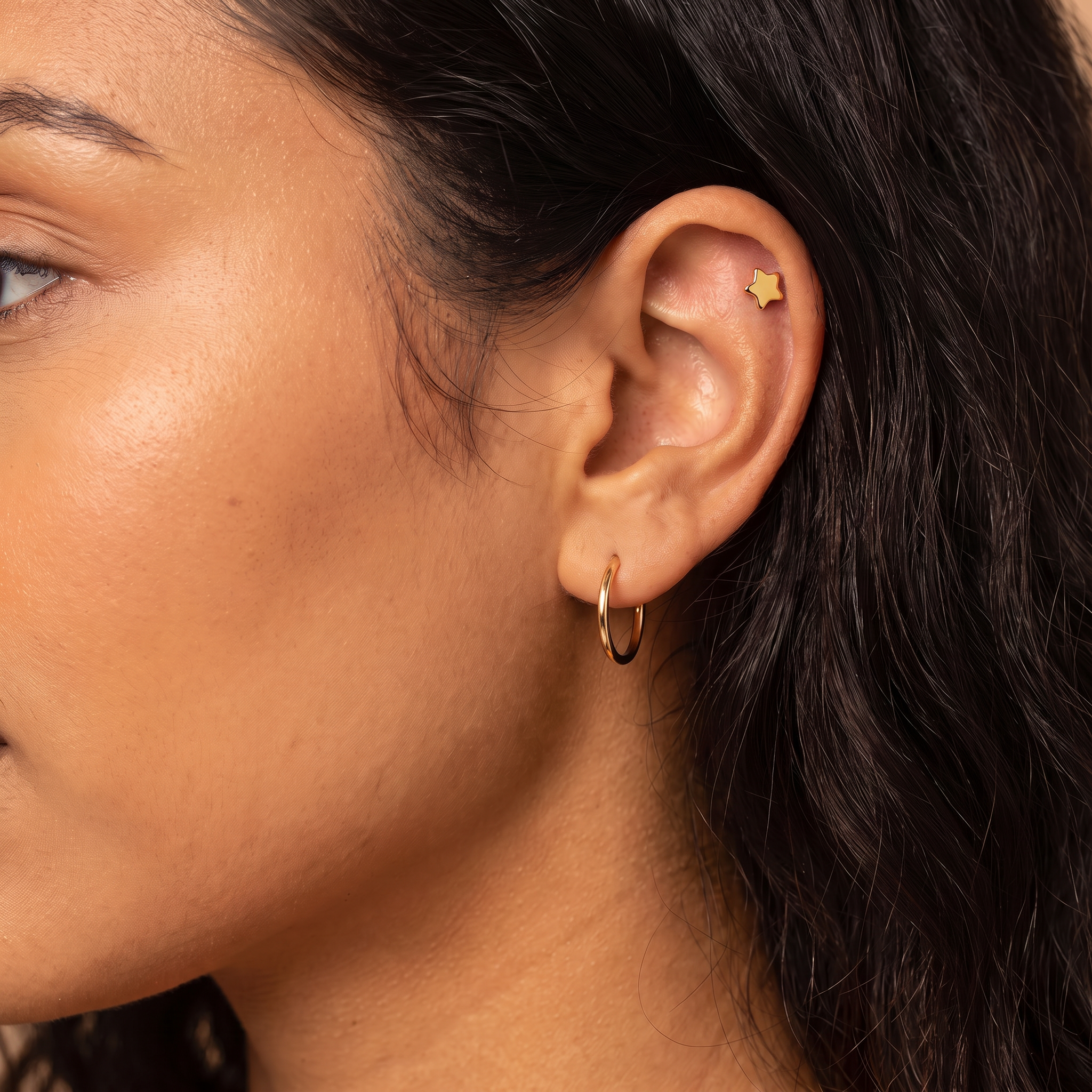 Close-up of a person's ear wearing a small gold hoop earring and a gold star-shaped cartilage piercing, with dark hair and warm skin tones.