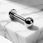 A polished stainless steel barbell piercing jewelry piece with spherical ends, resting diagonally on a white marble block with gray veining.