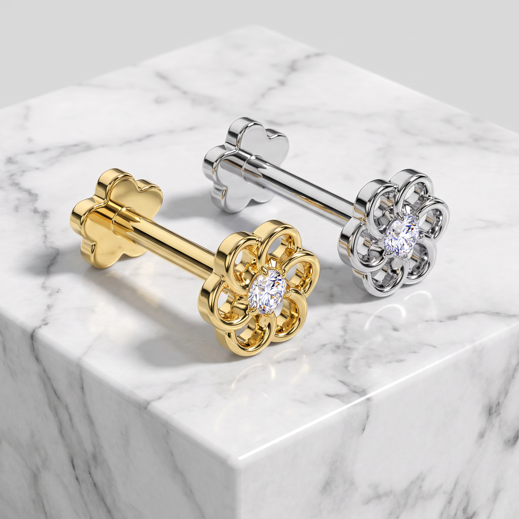 Two flower-shaped stud earrings displayed on a white marble block. One earring is yellow gold with a clear round gemstone, and the other is silver with a matching clear round gemstone.