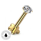 A gold barbell stud piercing with a clear faceted gemstone and a heart-shaped back, shown on a white background with a circular 'skin-safe waterproof' badge in the lower-left corner.