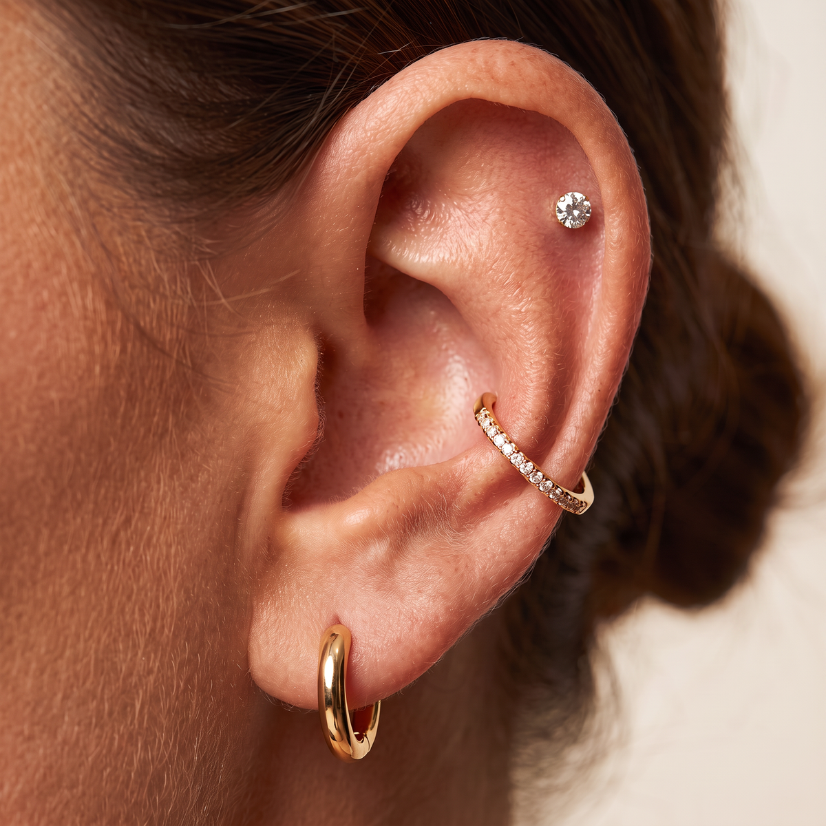 Close-up of a person's ear showcasing a three-piece gold ear cuff set: a small gold hoop in the lobe, a gold curved band with tiny clear stones along the front, and a single clear round gemstone stud in the lobe.