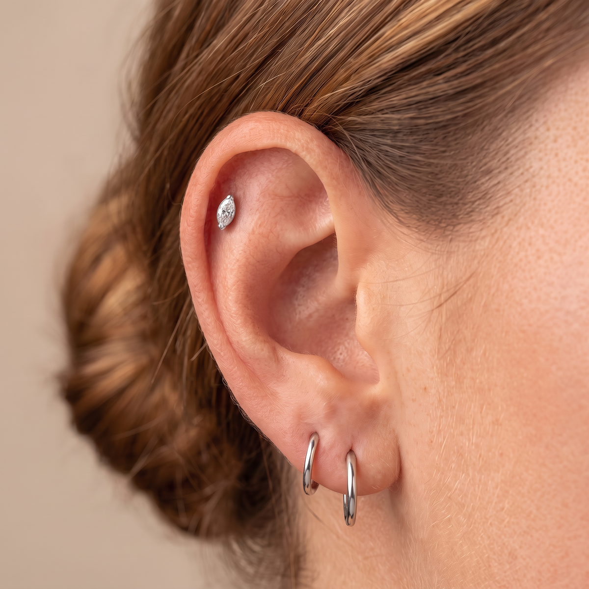 Close-up of a person's ear showcasing a multi-piece ear piercing set, including a small oval diamond stud on the upper lobe, two silver hoop earrings on the lower lobes, and a silver helix piercing.