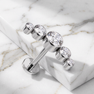 A silver-toned barbell-style body jewelry piece with five clear faceted gemstones of varying sizes, displayed on a white marble slab over a marble surface.