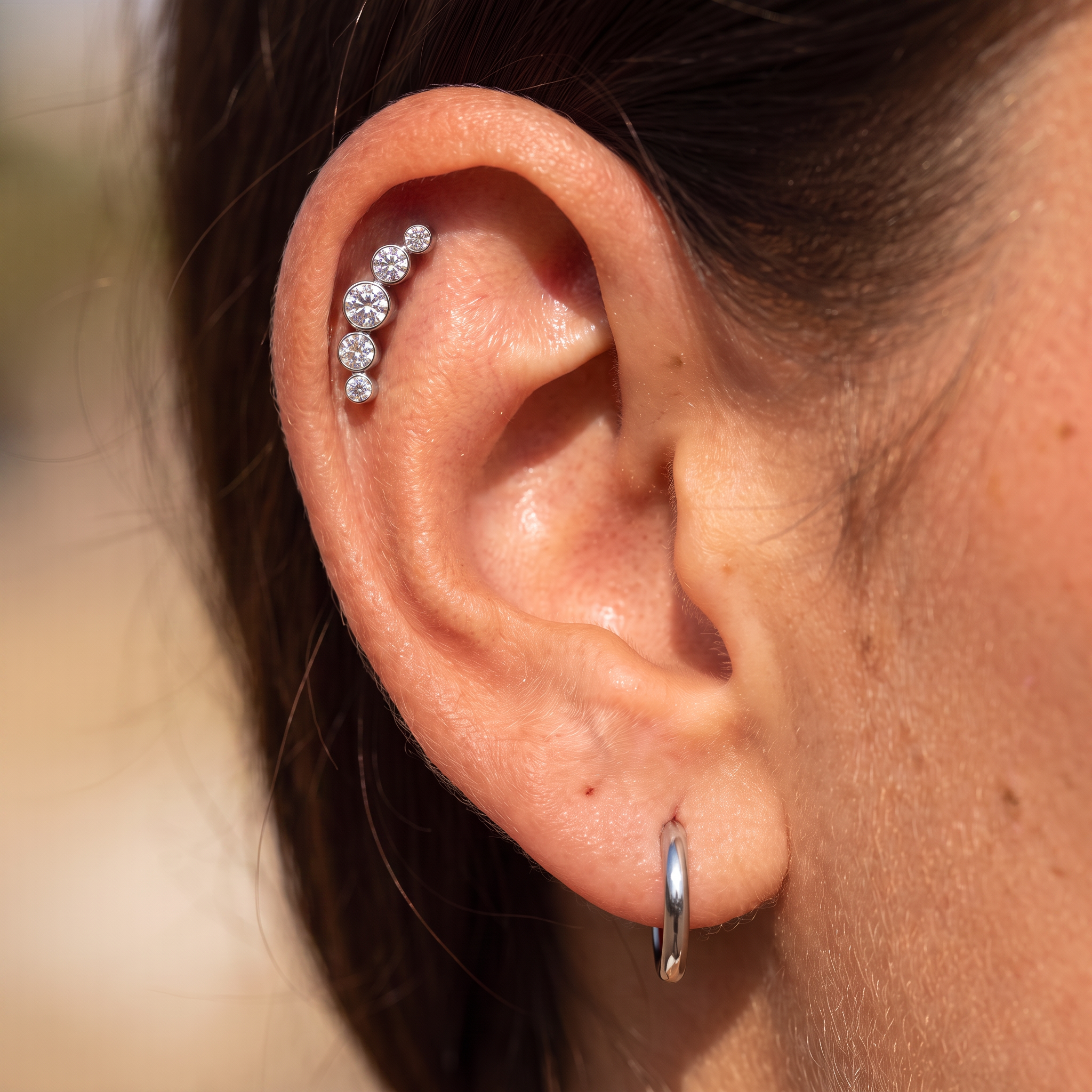 A close-up of a person's ear showcasing a silver ear cuff adorned with five clear, round-cut gemstones. A simple silver hoop earring is also visible on the lobe.