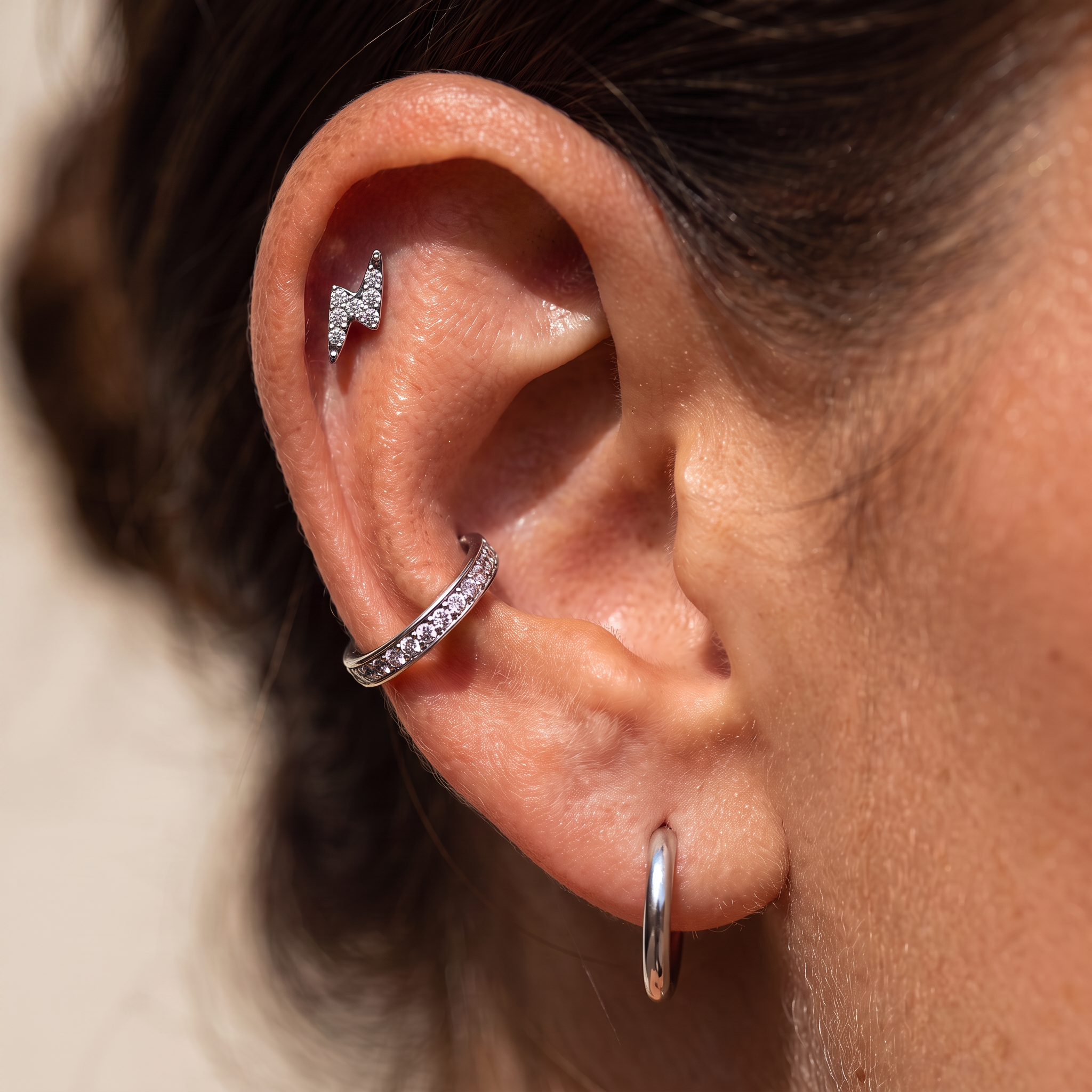 A close-up of a person's ear showcasing multiple silver ear piercings: a small lightning bolt stud in the upper helix, a pavé huggie hoop in the lobe, and a simple silver hoop earring in the lobe.